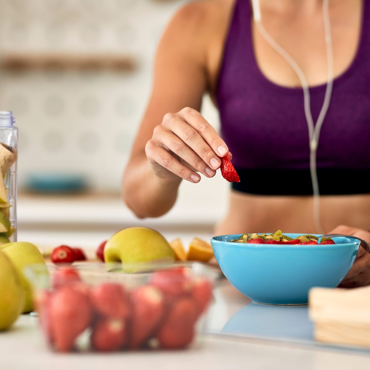 Close-up of athletic woman adding strawberries while making fruit salad in the kitchen.