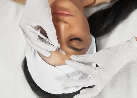 From above view of hyaluronic injection in female forehead. Cosmetologist using syringe with filler while brunette patient with closed eyes, in towel on head lying. Concept of cosmetology, beauty.