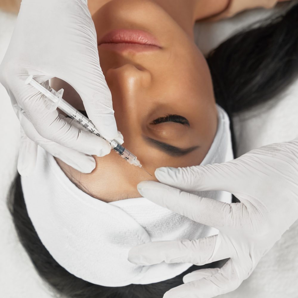From above view of hyaluronic injection in female forehead. Cosmetologist using syringe with filler while brunette patient with closed eyes, in towel on head lying. Concept of cosmetology, beauty.